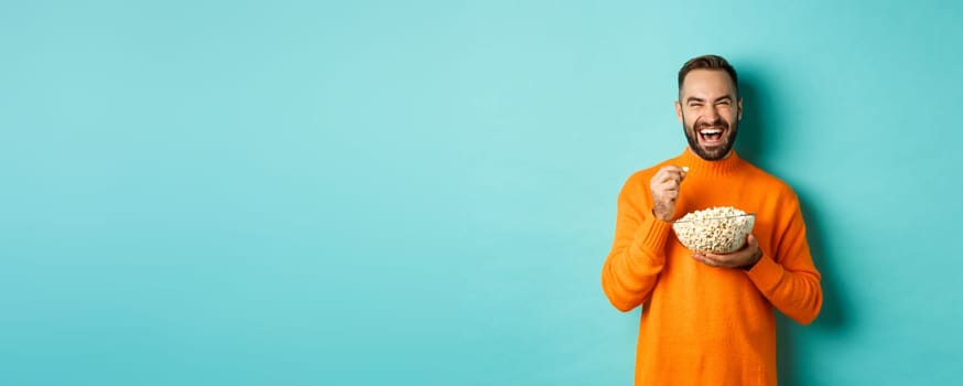 Excited young man watching interesting movie on tv screen, eating popcorn and looking amazed, blue background.