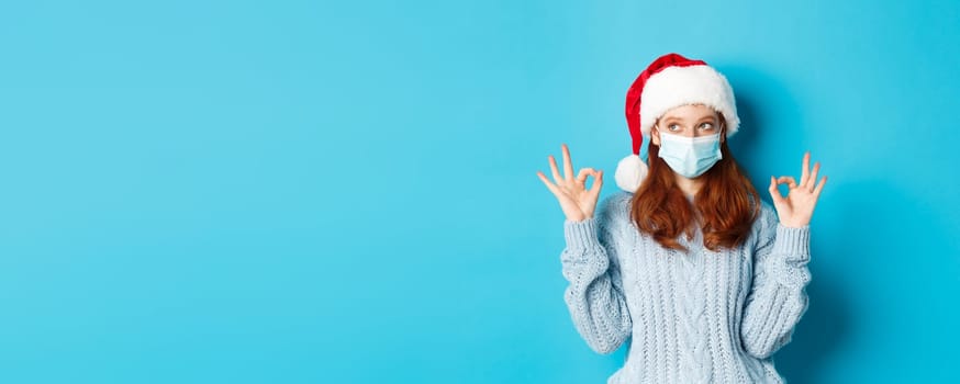 Christmas, quarantine and covid-19 concept. Cute teen redhead girl in santa hat and sweater, wearing face mask from coronavirus, showing okay signs, approve and praise something.