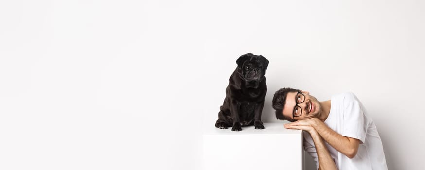 Handsome young man lay head near cute black pug, smiling and looking up at copy space, white background.