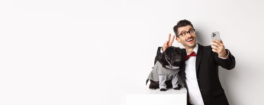 Cheerful dog owner in suit celebrating Christmas with dog, taking selfie on smartphone near cute black pug in costume, white background.