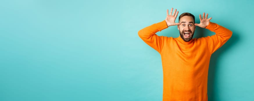 Image of handsome caucasian man making funny faces, mocking someone and smiling, standing against light blue background.