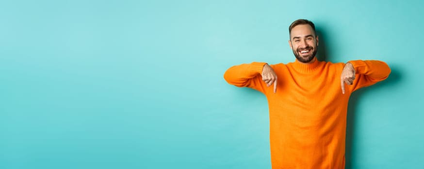 Happy adult man with beard pointing fingers down, smiling cheerful, showing advertisement, standing over turquoise background.