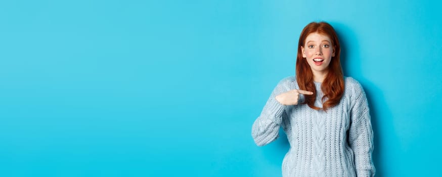 Hopeful redhead girl pointing at herself, standing over blue background.