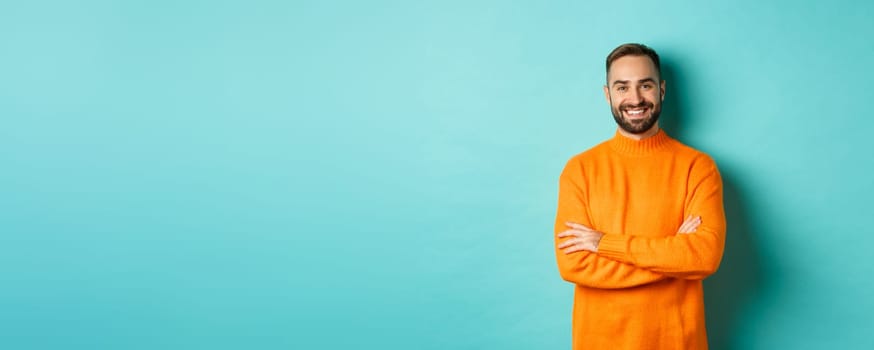 Handsome bearded male model in orange sweater, looking confident and smiling, cross arms on chest, standing over turquoise background.