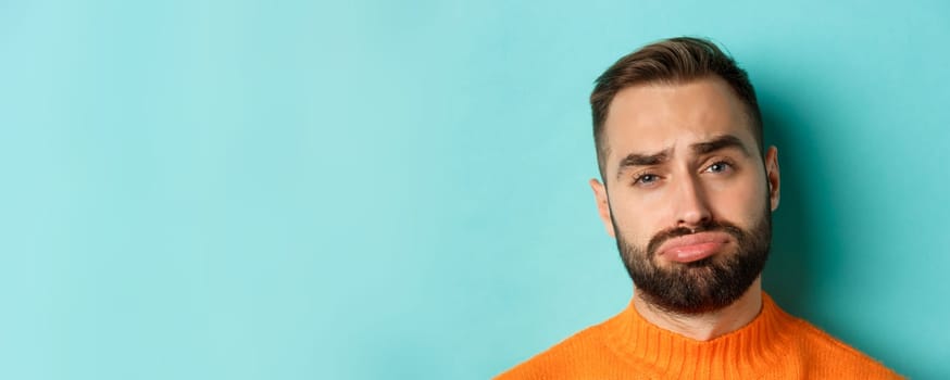 Headshot of sad and gloomy man complaining, pouting and frowning disappointed, standing against turquoise background.