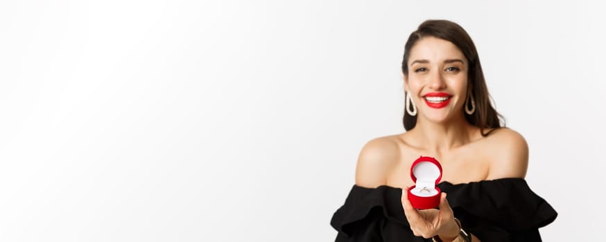 Fashion and beauty concept. Close-up of gorgeous brunette woman with red lips, black dress, showing engagement ring in box and smiling, getting married, standing over white background.