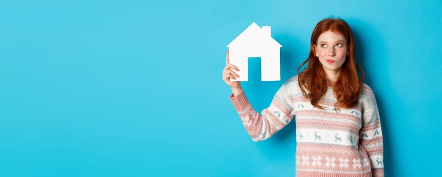 Real estate concept. Image of thoughtful redhead girl showing paper house model and thinking, searching for home or flat, standing against blue background.