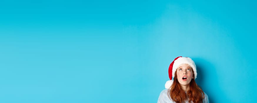 Winter holidays and Christmas eve concept. Head of pretty redhead girl in santa hat, appear from bottom and looking up at logo, seeing promo offer amazed, blue background.