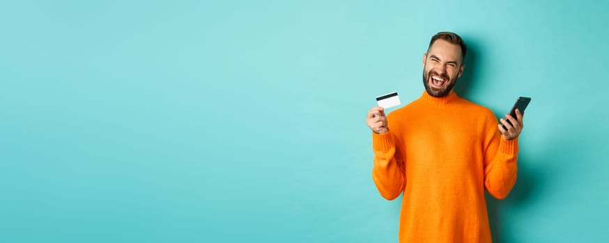 Online shopping. Happy young man using mobile phone and credit card, paying internet, light blue background.