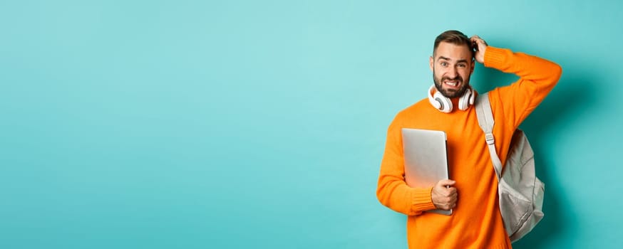 Confused male student looking puzzled and scratching head, holding laptop and backpack, standing over light blue background.