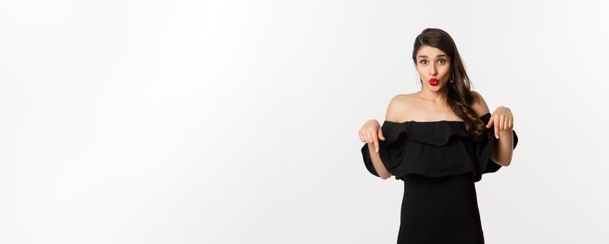 Fashion and beauty. Elegant woman in black dress pointing fingers down, showing promo and smiling, standing over white background.