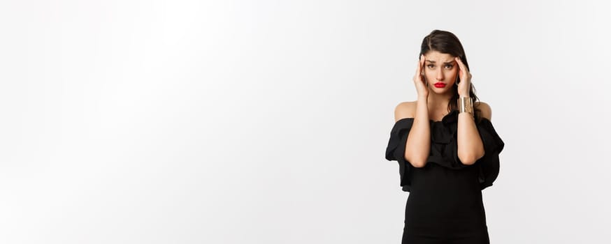 Fashion and beauty. Young modern woman in glamour dress, jewelry and makeup, touching head and looking exhausted, feeling dizzy, standing over white background.