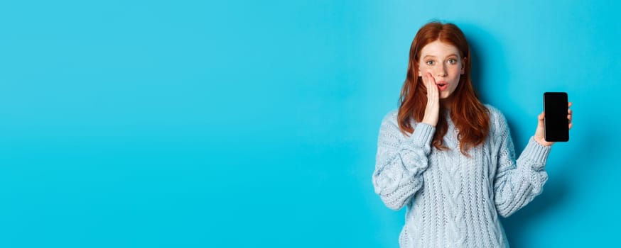 Amazed redhead girl looking at camera, showing smartphone screen, demonstrating online offer, standing over blue background.