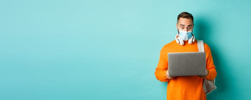 Happy young man in medical mask, working on laptop, looking shocked at computer screen, standing over light blue background.