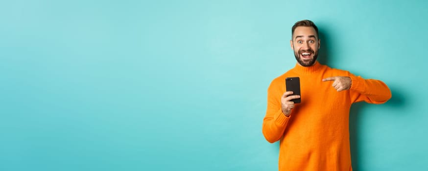 Excited man pointing at mobile phone, showing internet promo, standing over turquoise background, concept of online shopping.