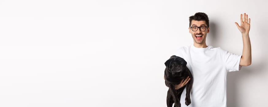 Handsome young man in glasses holding his black pug and waving hand, guy saying hello while carry dog with one arm, standing over white background.