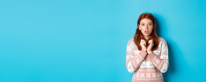 Close-up of cute redhead girl looking with anticipation and worry, staring at camera, standing in winter sweater against blue background.