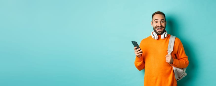 Caucasian man with headphones and backpack staring at camera amazed after reading phone notification, standing over turquoise background.