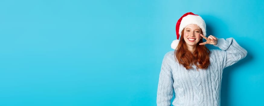 Winter holidays and Christmas Eve concept. Happy teenage girl with red hair, wearing santa hat, enjoying New Year, showing peace sign and smiling, standing over blue background.