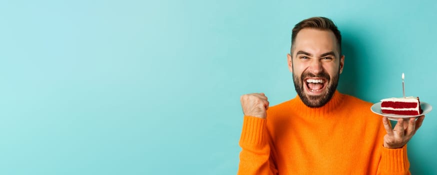 Close-up of happy adult man celebrating birthday, holding bday cake with candle and making wish, standing against turquoise background.