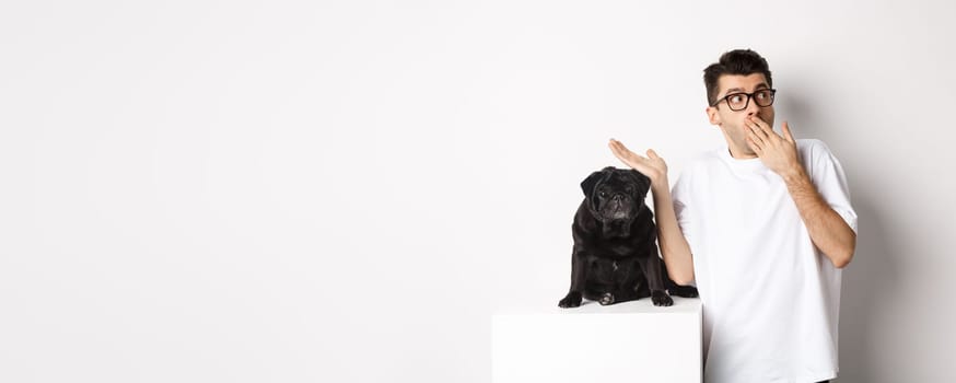 Surprised and shocked man in glasses, standing near cute black dog and staring right at logo, posing over white background.