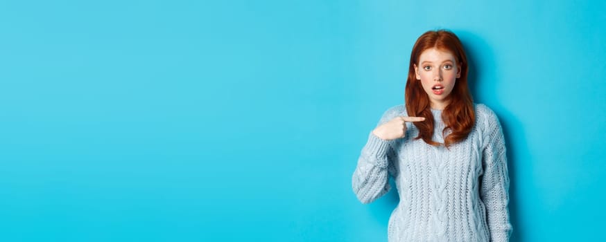 Confused redhead girl pointing at herself, being chosen, standing in sweater against blue background.