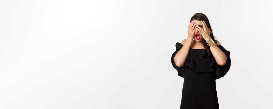 Fashion and beauty. Shocked young woman in black dress covering eyes, peeking through fingers at something embarrassing, cringe, standing over white background.