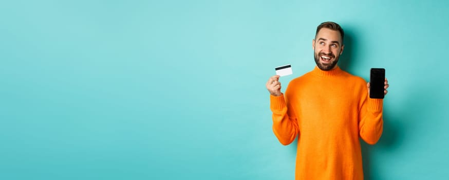 Online shopping. Thoughtful man shop in internet, showing mobile screen and credit card, looking at upper left corner and thinking, standing over light blue background.