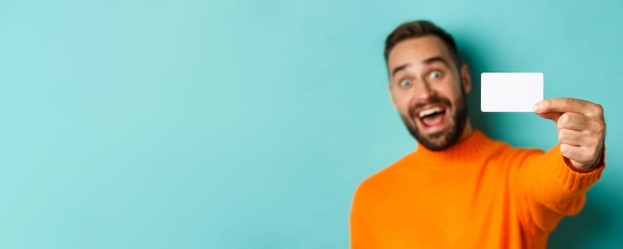 Close-up of handsome caucasian man going on shopping, showing credit card and smiling, standing over turquoise background.