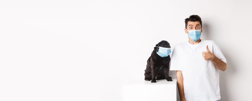 Covid-19, animals and quarantine concept. Image of funny young man and small dog in medical masks, owner showing thumb up in approval or like, white background.