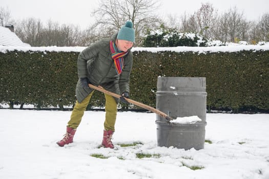 a middle-aged woman is collecting snow in a barrel with a shovel. For further watering plants in a greenhouse, the concept of protecting the environment and conserving natural resources