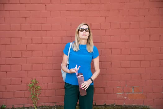 a blonde in a blue blouse against a brick wall drinks water from sports water bottle with time markers, to comply with the drinking regime, High quality photo