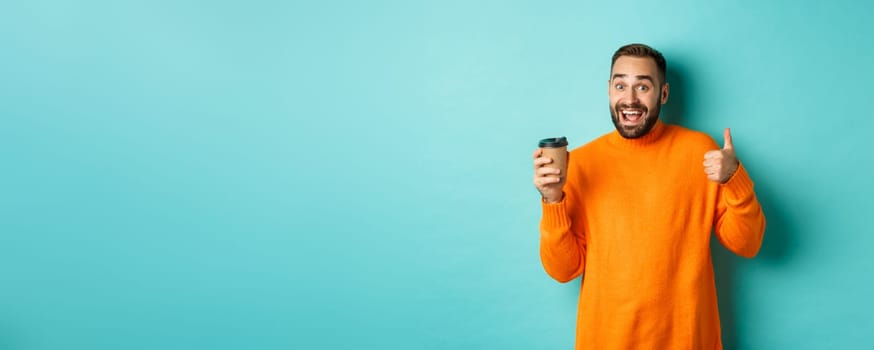 Handsome satisfied man showing thumbs-up, like coffee, smiling pleased, standing over turquoise background. Copy space