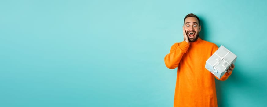 Holidays and celebration concept. Surprised man receiving gift, looking happy at present and smiling, standing over blue background.