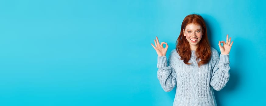 Confident redhead girl showing okay signs, praising choice, approve and agree, like something, standing over blue background. Copy space