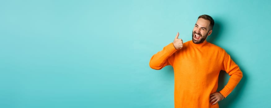 Happy satisfied male model showing thumb up, looking pleased at upper left corner logo, approve and recommend product, standing over light blue background.