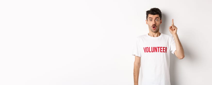 Handsome young man in volunteer t-shirt having an idea, raising finger and saying suggestion, pointing up, standing over white background.