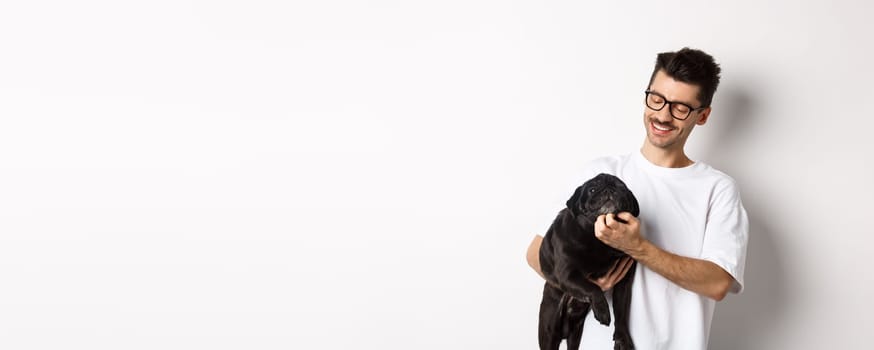 Happy dog owner scratching cute black pug face and smiling, pet his animal, standing over white background.