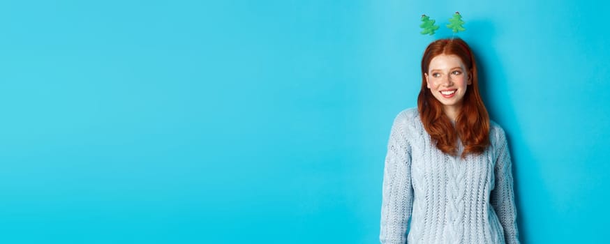 Winter holidays and Christmas sales concept. Cute redhead girl in funny New Year headband smiling, looking left at logo, standing over blue background.