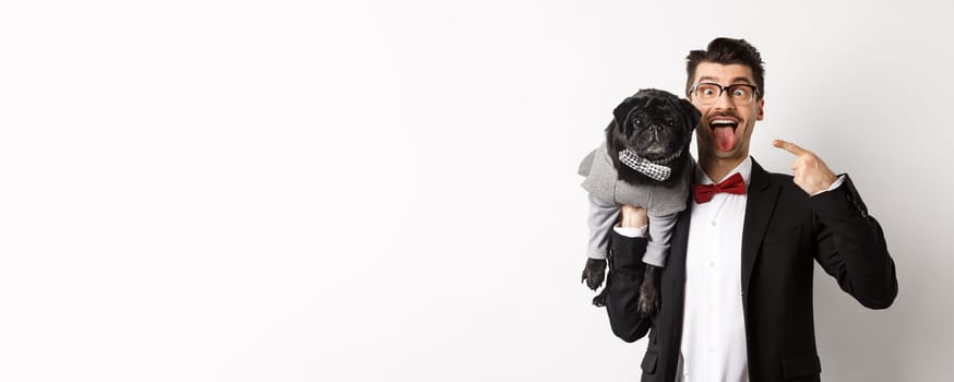 Funny young man in party suit, showing tongue and making grimaces, pointing at cute black dog in winter clothes, standing over white background.