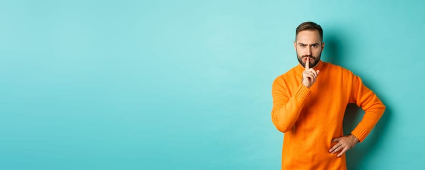 Angry bearded guy telling be quiet, hushing and frowning displeased, shut up gesture, standing over turquoise background.