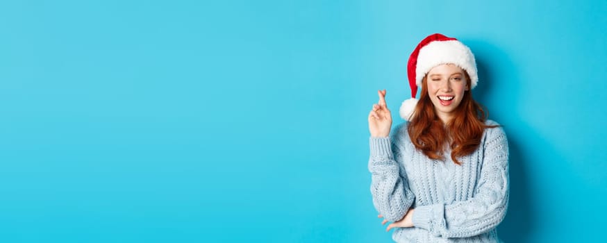Winter holidays and Christmas Eve concept. Hopeful redhead girl in Santa hat, making wish on xmas, cross fingers for good luck and winking, standing over blue background.