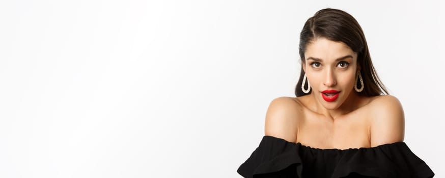 Close-up of gorgeous brunette woman wearing elegant earrings and black dress, looking sensual at camera with piercing gaze and opened mouth, standing over white background.