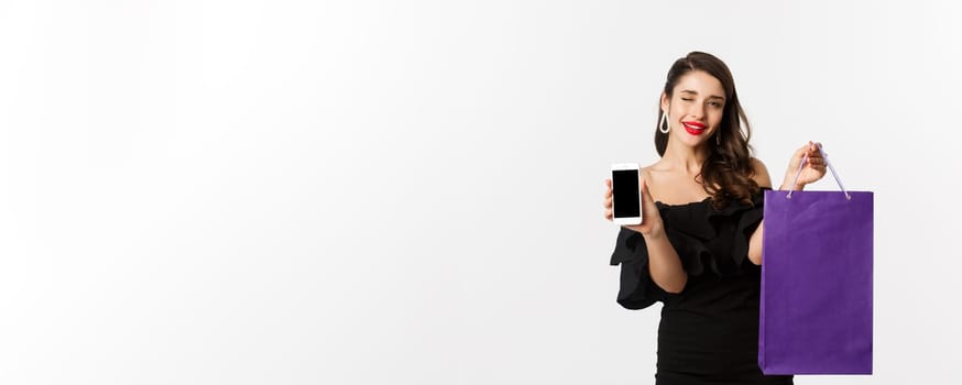 Beauty and shopping concept. Beautiful and stylish woman winking, showing smartphone screen and bag, buying online, standing over white background.