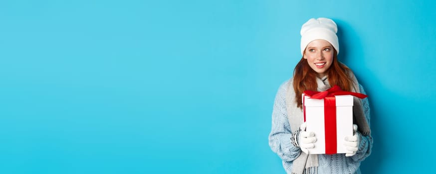 Winter holidays and Christmas sales concept. Thoughtful redhead woman standing in white beanie and gloves, holding holiday gift and looking left, thinking, standing over blue background.