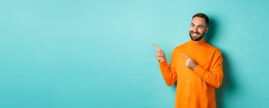 Attractive bearded male model in orange sweater, smiling satisfied and pointing fingers left, standing over turquoise background.