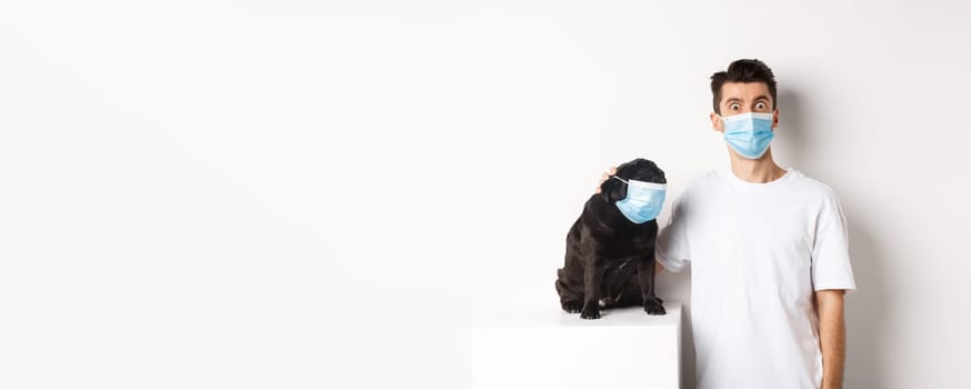 Covid-19, animals and quarantine concept. Image of funny young man and pug in medical masks, staring at camera, standing over white background.