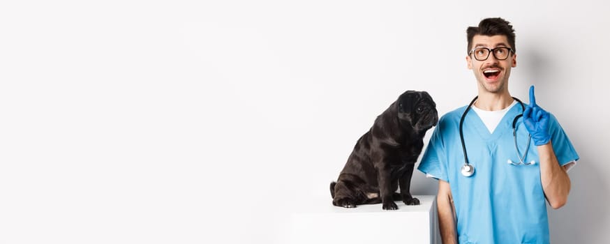 Handsome young doctor at vet clinic pointing finger up and looking amazed, standing near cute black pug dog, white background.