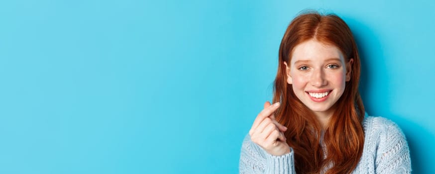 Headshot of cute caucasian woman with red hair and freckles showing heart sign and smiling, standing against blue background.