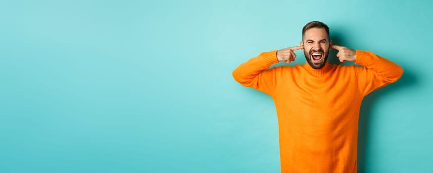 Displeased man shut ears and grimacing, loud neighbours, standing annoyed with noise, shouting angry and distressed, light blue background.